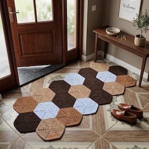 May include: A runner rug composed of interconnected hexagonal crochet motifs in shades of dark brown, light brown, tan, and white. The rug features a rectangular leather-like tag with the text "Mirena" stitched onto one of the tan hexagonal pieces.