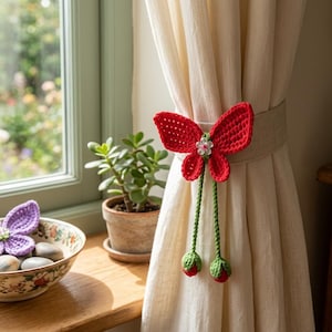 Könnte beinhalten: Ein roter gehäkelter Schmetterling-Gardinenhalter mit grünen Stielen und roten und grünen Knospen. Der Schmetterling ist an einem beigen Vorhang befestigt. Ein lila gehäkelter Schmetterling befindet sich in einer dekorativen Schale auf einer Holzfensterbank.