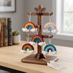 May include: A wooden display stand holding five crocheted rainbow keychains with white clouds at each end. The rainbows feature various color palettes including bright primary colors, pastels, and deep blues. A tag reads Handcrafted Crochet Keychains Set of 5.