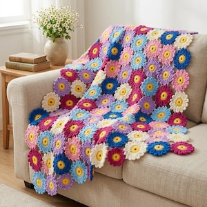 May include: A vibrant crochet blanket featuring daisy motifs in shades of pink, blue, yellow, purple, and white, draped over a beige sofa. A vase of flowers sits on a wooden side table. The blanket is a handmade item.