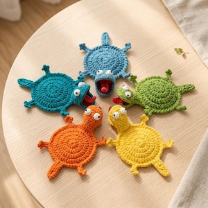 Pode incluir: Cinco bases para copos em croché com formato de criaturas de desenhos animados em azul, azul-petróleo, verde, laranja e amarelo. Cada base tem um corpo circular, quatro membros, uma cauda e uma cabeça com dois olhos brancos. Três têm bocas abertas com línguas vermelhas.