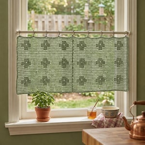 May include: A sage green crocheted window curtain with a floral pattern hangs from a wooden rod. The curtain is made of a mesh-like material. A small potted plant sits on the windowsill below the curtain.