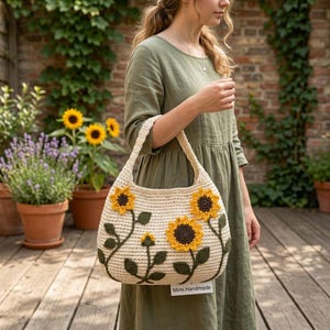 May include: A cream-coloured crochet shoulder bag featuring three yellow sunflower appliqués with dark brown centres and dark green stems and leaves. A small white rectangular tag with the black text Mimi Handmade is attached to the bottom.