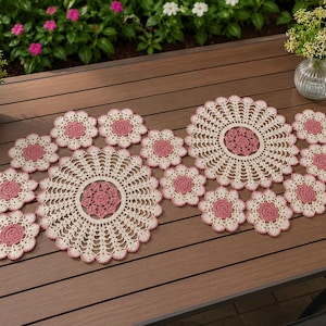 May include: A collection of cream and dusty rose crochet doilies arranged in a cluster. The set includes two large circular doilies with intricate openwork patterns and several smaller matching floral-shaped doilies with dark pink centers.
