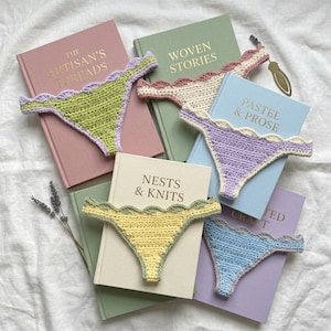 May include: Five crocheted thong-style garments in lime green, cream, lavender, yellow, and light blue, each featuring a scalloped edge. These items are arranged on top of five hardcover books titled The Artisan's Threads, Woven Stories, Pastel & Prose, Nests & Knits, and a partially visible book, accompanied by a sprig of dried lavender.