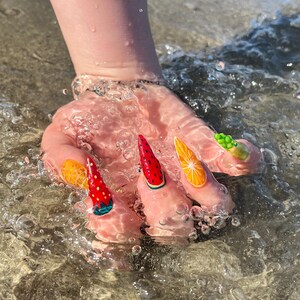 May include: A hand with long, decorated fingernails in the water. The nails feature colorful fruit designs, including a strawberry, watermelon, and orange slices. The water is clear and the background is a sandy beach.