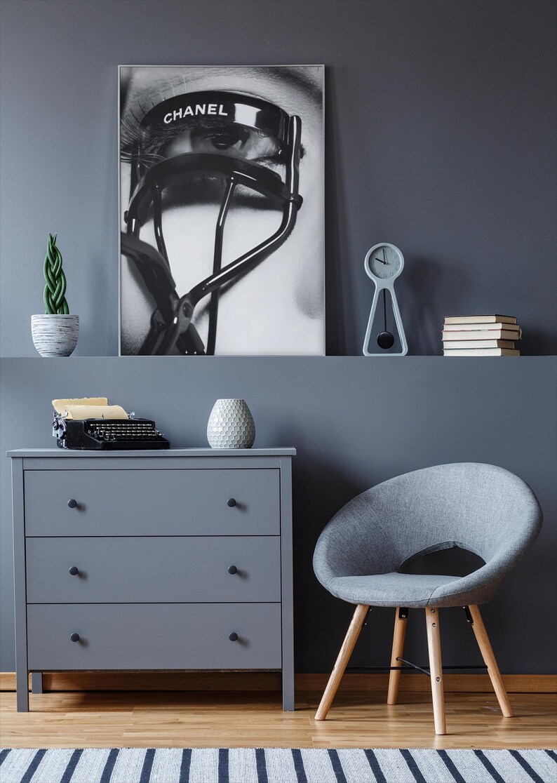 Puede incluir: Una impresi&oacute;n en escala de grises de un ojo con un rizador de pesta&ntilde;as CHANEL se muestra sobre una c&oacute;moda gris. Un peque&ntilde;o reloj, libros y una planta en maceta est&aacute;n en el estante. Una silla gris y una m&aacute;quina de escribir vintage tambi&eacute;n est&aacute;n en la habitaci&oacute;n.