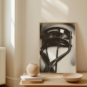 Puede incluir: Impresi&oacute;n enmarcada en blanco y negro que presenta un primer plano de un ojo con largas pesta&ntilde;as y un rizador de pesta&ntilde;as Chanel. La palabra "CHANEL" est&aacute; impresa en el rizador. La impresi&oacute;n se muestra en una pared de color claro, con una peque&ntilde;a mesa y objetos decorativos en primer plano.
