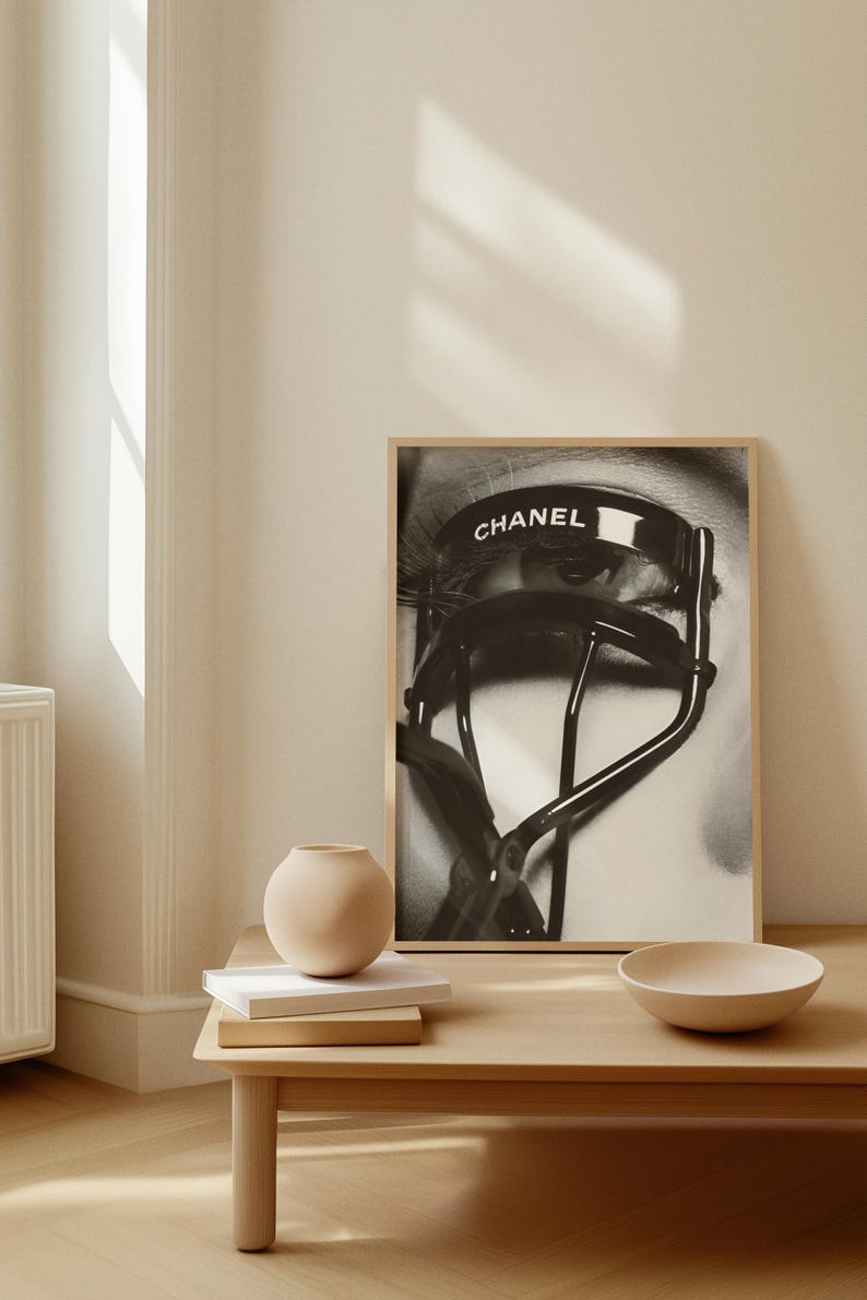 Puede incluir: Impresi&oacute;n enmarcada en blanco y negro que presenta un primer plano de un ojo con largas pesta&ntilde;as y un rizador de pesta&ntilde;as Chanel. La palabra "CHANEL" est&aacute; impresa en el rizador. La impresi&oacute;n se muestra en una pared de color claro, con una peque&ntilde;a mesa y objetos decorativos en primer plano.