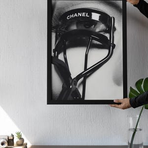Puede incluir: Impresi&oacute;n enmarcada en blanco y negro que presenta un primer plano de un ojo con largas pesta&ntilde;as y un rizador de pesta&ntilde;as CHANEL. La imagen est&aacute; sostenida por una persona. La impresi&oacute;n se muestra en un escritorio de madera.