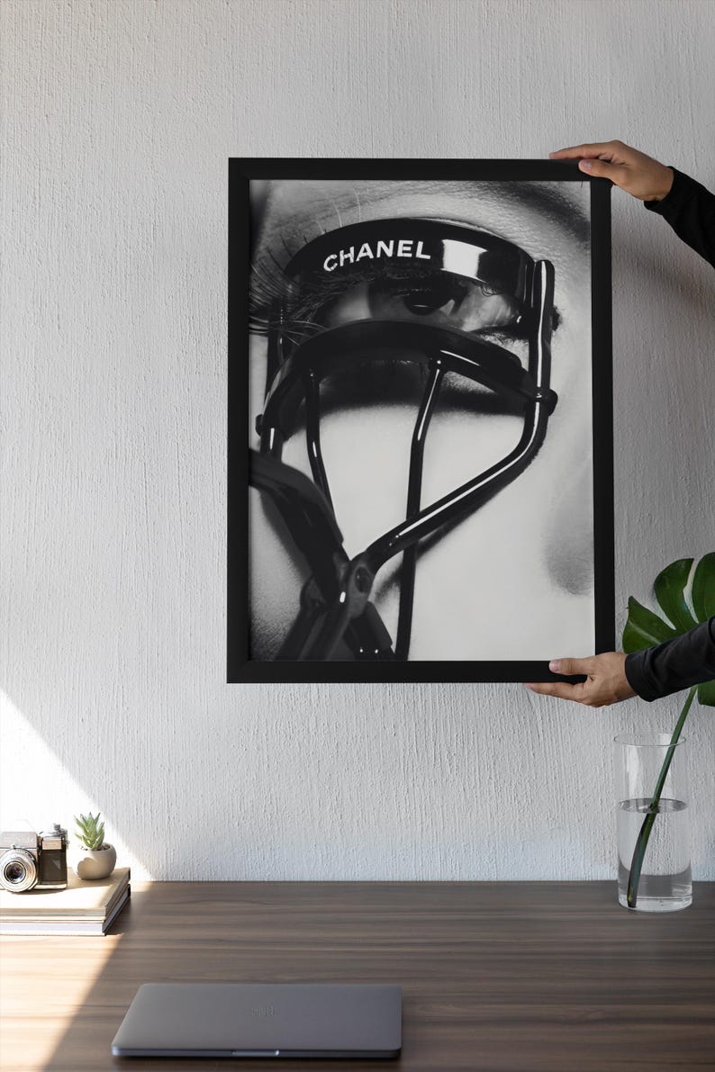 Puede incluir: Impresi&oacute;n enmarcada en blanco y negro que presenta un primer plano de un ojo con largas pesta&ntilde;as y un rizador de pesta&ntilde;as CHANEL. La imagen est&aacute; sostenida por una persona. La impresi&oacute;n se muestra en un escritorio de madera.