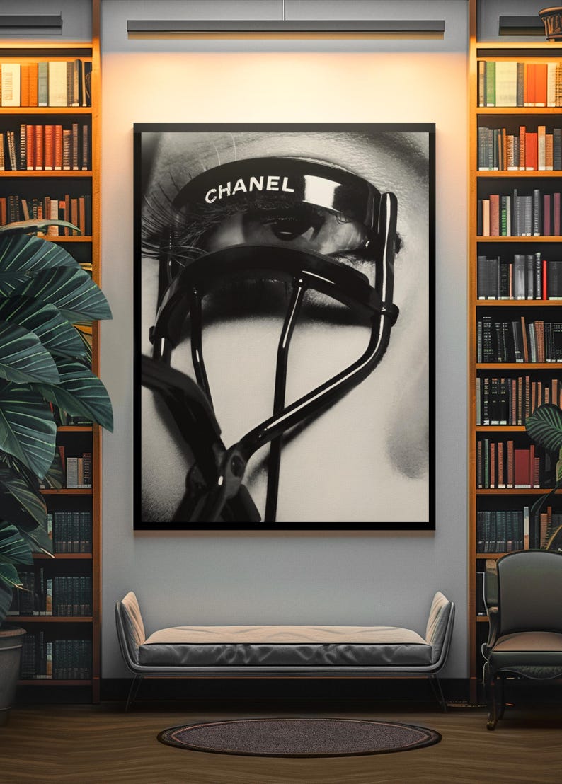 Puede incluir: Impresi&oacute;n en blanco y negro enmarcada que presenta un primer plano de un ojo con largas pesta&ntilde;as y un rizador de pesta&ntilde;as Chanel. La impresi&oacute;n se exhibe en una habitaci&oacute;n con estanter&iacute;as, un banco y una silla.