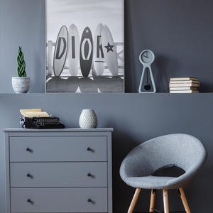 May include: A gray dresser with three drawers, a gray chair, and a black and white framed print of surfboards with the word "DIOR" on them. A small clock, books, and a decorative plant are on a shelf above the dresser.