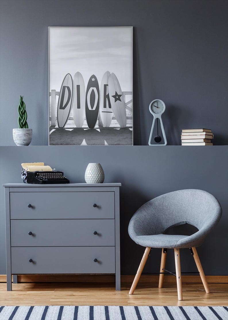May include: A gray dresser with three drawers, a gray chair, and a black and white framed print of surfboards with the word "DIOR" on them. A small clock, books, and a decorative plant are on a shelf above the dresser.
