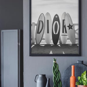 May include: Black and white framed print featuring five surfboards with the letters "DIOR" on them. The surfboards are leaning against a white fence on a beach. Below the frame are decorative items, including vases and a plant.