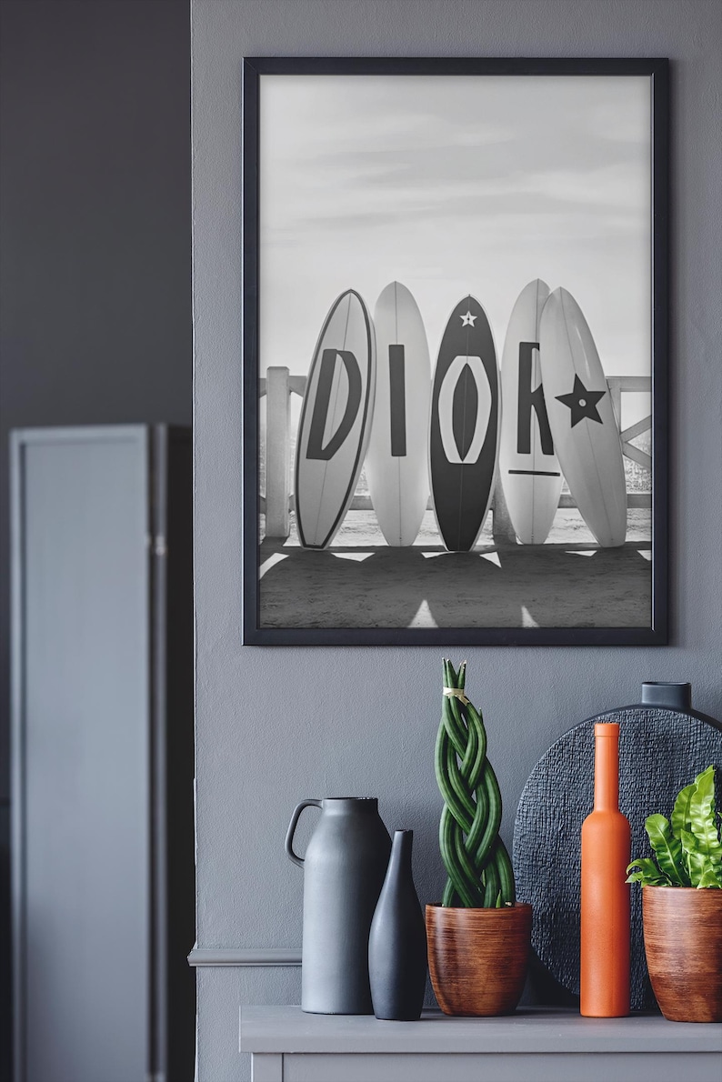 May include: Black and white framed print featuring five surfboards with the letters "DIOR" on them. The surfboards are leaning against a white fence on a beach. Below the frame are decorative items, including vases and a plant.