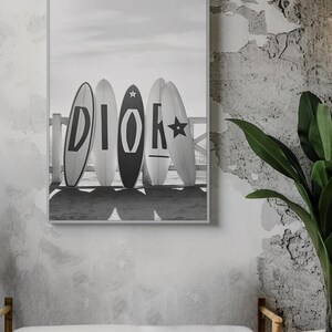 May include: Black and white framed print featuring five surfboards with the word "DIOR" spelled out in large letters. The surfboards are leaning against a wooden fence on a beach, with a cloudy sky in the background.