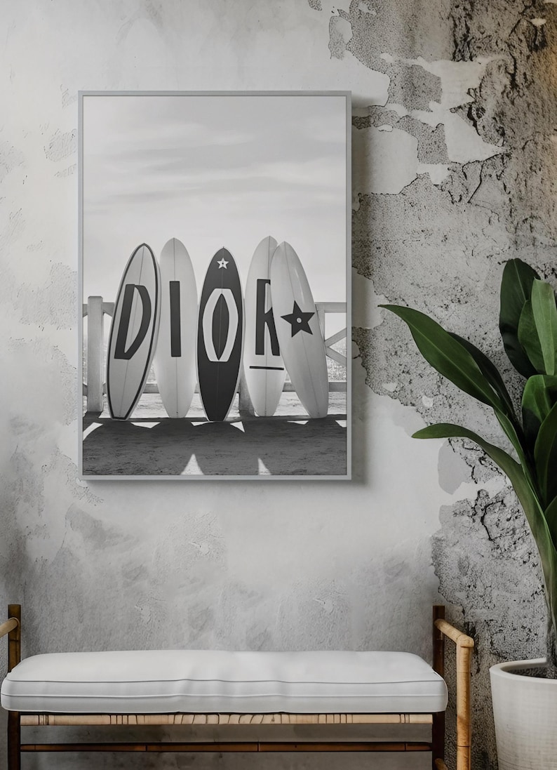 May include: Black and white framed print featuring five surfboards with the word "DIOR" spelled out in large letters. The surfboards are leaning against a wooden fence on a beach, with a cloudy sky in the background.