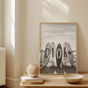 May include: A framed black and white print featuring five surfboards with the word "DIOR" on them. The surfboards are leaning against a wooden fence on a beach. A small, round vase and a bowl sit on a wooden table.