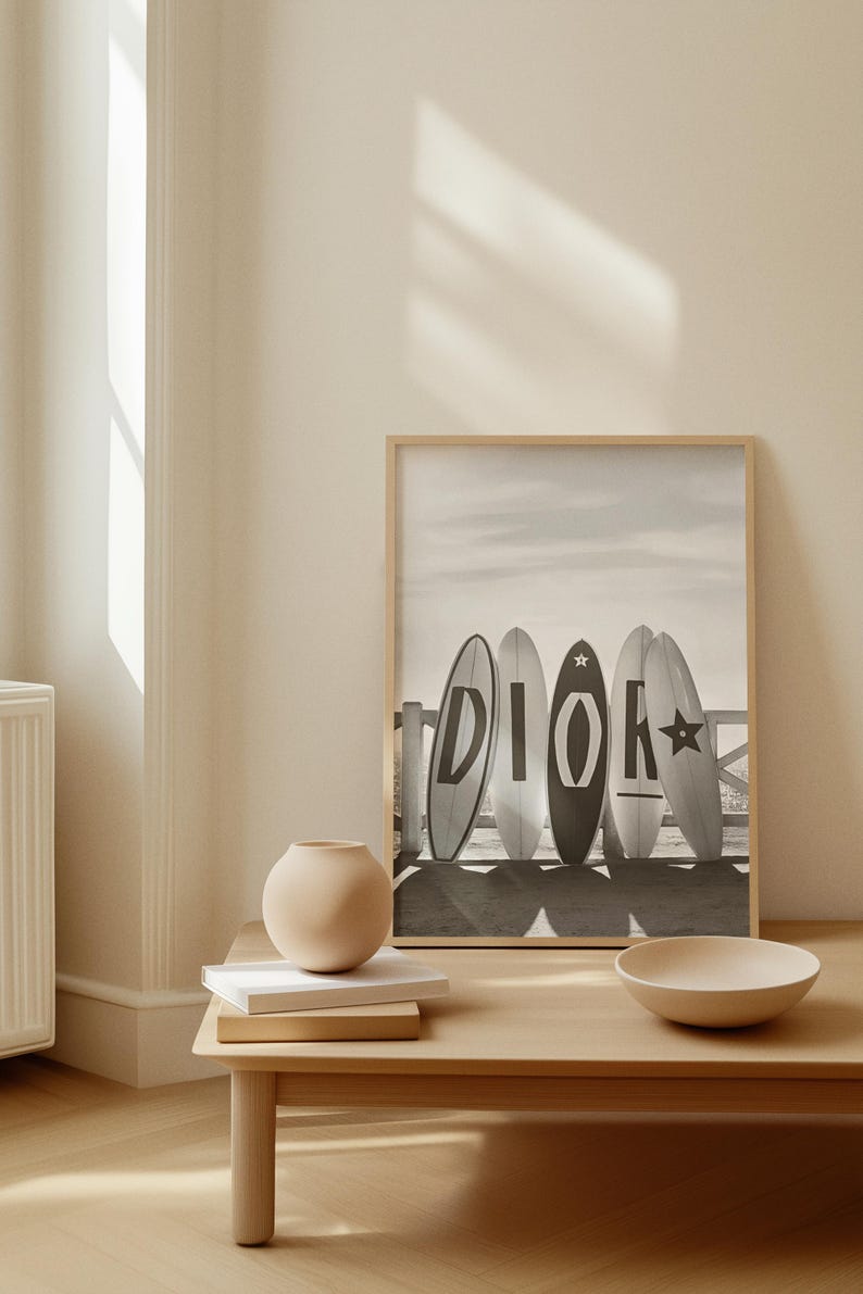 May include: A framed black and white print featuring five surfboards with the word "DIOR" on them. The surfboards are leaning against a wooden fence on a beach. A small, round vase and a bowl sit on a wooden table.