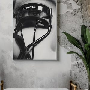 Puede incluir: Impresi&oacute;n enmarcada en blanco y negro que presenta un primer plano de un ojo con largas pesta&ntilde;as y un rizador de pesta&ntilde;as negro. La palabra "CHANEL" est&aacute; impresa en el rizador. La impresi&oacute;n se muestra en una pared gris texturizada.