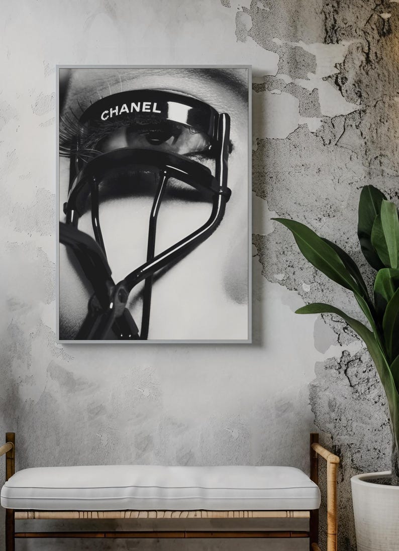 Puede incluir: Impresi&oacute;n enmarcada en blanco y negro que presenta un primer plano de un ojo con largas pesta&ntilde;as y un rizador de pesta&ntilde;as negro. La palabra "CHANEL" est&aacute; impresa en el rizador. La impresi&oacute;n se muestra en una pared gris texturizada.