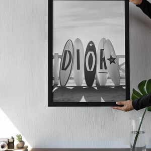 May include: Black and white framed print featuring five surfboards leaning against a white fence on a beach. The surfboards display the letters "DIOR" in a bold, stylized font. The image is held by a person.