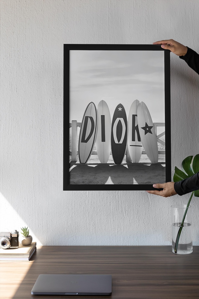 May include: Black and white framed print featuring five surfboards leaning against a white fence on a beach. The surfboards display the letters "DIOR" in a bold, stylized font. The image is held by a person.