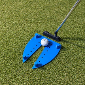 May include: A blue plastic golf putting training aid with four vertical pegs and a series of alignment holes. A white Callaway golf ball sits between the two arms of the device, positioned in front of a black metal putter head.