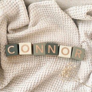 May include: Six wooden blocks arranged in a row spelling the name CONNOR. The blocks alternate between a dark sage green and a cream colour, each featuring a single capital letter in a natural wood finish engraved on the front face of the cube.