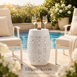 May include: A white ceramic garden stool with a cylindrical shape and intricate cut-out floral and paisley patterns. A glass wine bottle and two clear stemmed wine glasses are placed on the flat top surface of the stool between two chairs.