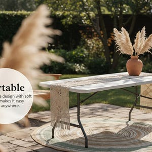 May include: A white folding table with black legs is set up outdoors. A decorative runner and vase with dried flowers sit on top. The table is on a round rug. The image includes the word "Portable".