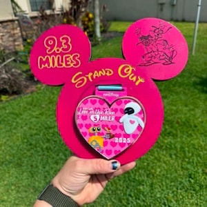 May include: A pink Mickey Mouse-shaped medal holder with the text "9.3 MILES" and "Stand Out". A heart-shaped medal with the text "Love on the Run 5 Miler" and images of WALL-E and Eve is displayed. The year "2025" is also visible.