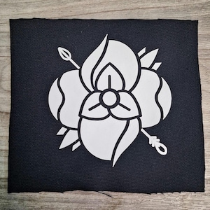 May include: A square, black fabric patch featuring a white floral design. The design includes a stylized flower with petals, leaves, and two arrows. The patch is likely for sewing or ironing onto clothing or accessories.