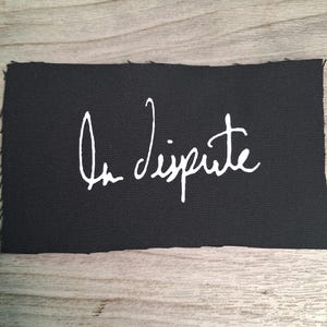 May include: A black rectangular patch with the handwritten text "In Dispute" in white cursive script. The edges of the patch are unfinished, with visible frayed threads. The patch is likely made of a durable fabric.