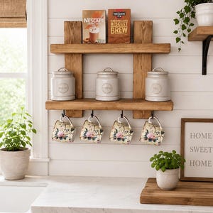May include: A two-tier wooden wall shelf with vertical supports, holding three light grey ceramic canisters labelled Sugar, Coffee, and Tea. Four floral patterned mugs hang from black metal hooks beneath the bottom shelf. Two boxes of Nescafe and Yorkshire Tea sit on the top shelf.