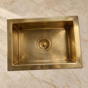May include: A rectangular kitchen sink crafted from brushed brass with a metallic gold finish. The sink features a centred circular drain assembly with a matching brass strainer, showcasing a smooth, polished surface with subtle water droplets.