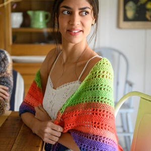 May include: A colorful, handmade crochet shawl in a rainbow of colors: purple, blue, green, orange, and red. The shawl is draped over the shoulders. The open weave design is visible. The shawl is a cozy accessory.