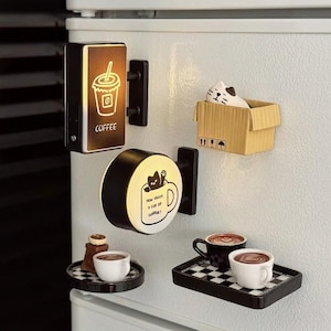Puede incluir: Un conjunto de imanes para nevera con temática de café. Incluye un letrero con un gráfico de taza de café y la palabra "COFFEE", un letrero redondo con un gato en una taza, un gato en una caja de cartón y tazas de café en miniatura en bandejas.