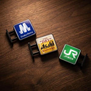 Może przedstawiać: Trzy kwadratowe metalowe znaki z czarnymi wspornikami. Znaki przedstawiają logo Osaka Metro (niebieskie i białe), piwa Asahi Super Dry (żółte i białe) oraz JR East Japan (zielone i białe). Znajdują się na drewnianej powierzchni.