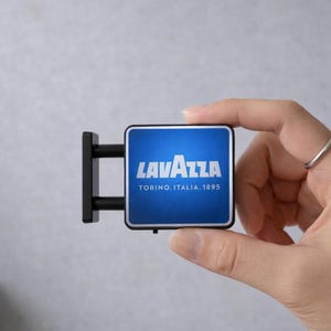 Może przedstawiać: Miniaturowy szyld Lavazza z niebieskim tłem i białym tekstem. Na szyldzie widnieje napis "LAVAZZA TORINO, ITALIA, 1895". Szyld jest trzymany w dłoni i ma czarny wspornik montażowy.