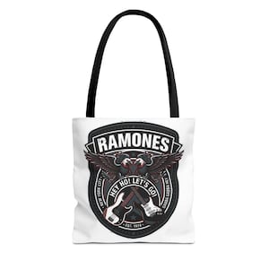 Pode incluir: Bolsa tote branca com alças pretas. O design apresenta a palavra "RAMONES", duas águias, duas guitarras e os textos "HEY HO! LET'S GO!" e "GABBA GABBA HEY". O texto "EST. 1974" também é visível.