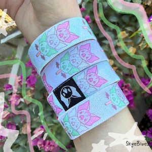 May include: A set of four light blue elastic wristbands featuring a repeating pattern of pink and green bats. The bands are stacked together, and one band displays a small black rectangular tag with a white bat icon graphic.