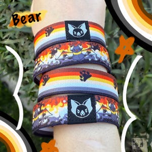 May include: Four colorful wristbands are stacked on top of each other. Each wristband has a different design, including a rainbow pattern, a bat, and paw prints. The word "Bear" is written in orange on the top left.