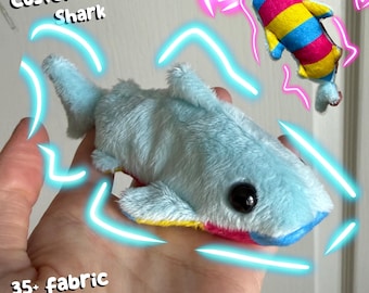May include: A plush shark toy with light blue fur, black eyes, and a multi-colored underside featuring pink, blue, and yellow stripes. The image includes the text Custom Fidget Shark and 35+ fabric options :3 with glowing neon blue and pink accents.