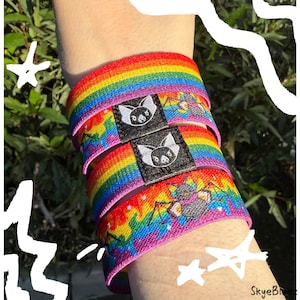 May include: Rainbow-striped elastic wristbands with bat and star graphics. Each band features a black label with a white bat design. The bands are stacked on a wrist, showcasing the vibrant colors and playful design.