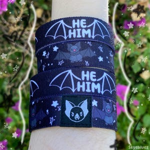 May include: Black wristbands with a bat and star pattern. The text "HE HIM" is printed in white on the bands, along with a cartoon bat illustration. The bands are worn on a wrist.