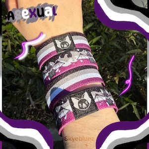 May include: Asexual pride wristbands in black, grey, white, and pink. The bands feature a bat logo and the word "Asexual" in purple. The wristbands are stacked on a wrist.