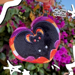 May include: A heart-shaped plush keychain featuring a black bat embroidery with red eyes. The plush has a gradient of orange, pink, and purple colors. A red plastic clip is attached to the top. The text 40+ flag options is visible.