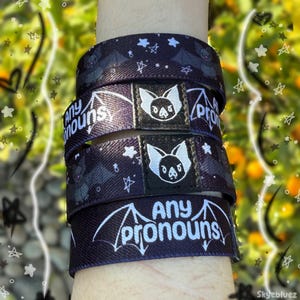 May include: Dark blue wristbands with a bat and star pattern. Each band features a white label with a bat design and the words "Any pronouns" in white. The bands are stacked on top of each other.